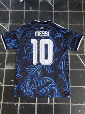 ARGENTINA VM TRÖJA | MESSI#10 - Snygg Argentina fotbollströja från Adidas med Messi och nummer 10 på ryggen. Tröjan är svart med blåa och ljusblåa mönster, vita detaljer och klassiska Adidas-ränder på axlarna. Perfekt för dig som älskar fotboll och vill sticka ut på planen.