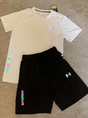 Under Armour träningsset svart/vit - Snyggt träningsset från Under Armour med en vit t-shirt och svarta shorts. Setet har regnbågsfärgade loggor och textdetaljer. Tillverkat i ett lätt och snabbtorkande material som passar perfekt till träning och sport.