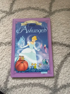 Askungen Disney klassiker bok - Säljer boken 'Askungen' från serien Våra bästa klassiker av Disney. Boken har ett lila hårt omslag med illustration av Askungen, pumpa och möss på framsidan. Perfekt för dig som gillar sagor och klassiska berättelser. Utgiven av Egmont Kids Media.