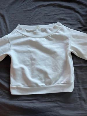 Vit sweatshirt med bred krage - En stilren vit sweatshirt med bred ribbad krage och muddar vid ärmslut och nederkant. Tröjan har en enkel och clean design utan tryck eller logga, perfekt för en avslappnad look. Tillverkad i mjukt bomullsmaterial som känns skönt mot huden.