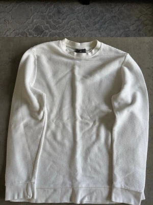 Vit ribbad sweatshirt från H&M - Vit långärmad sweatshirt från H&M med ribbad struktur och rund halsringning. Tröjan har en avslappnad passform och är tillverkad i ett mjukt material som känns skönt mot huden. Perfekt för en clean och stilren look.