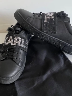 Svarta sneakers från Karl Lagerfeld - Svarta sneakers i läder-look från Karl Lagerfeld med bred kardborre över vristen dekorerad med glittrande stenar och texten 'KARL'. Är i bra skick och har inte anvönt dom på länge!😊