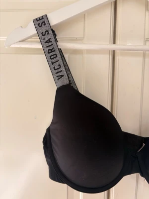 Svart push-up bh Victoria's Secret - Snygg svart push-up bh från Victoria's Secret med vadderade kupor och breda axelband med logotyp i silver. Axelbanden är justerbara och bh:n har en klassisk hakknäppning bak. Materialet känns mjukt och stretchigt för extra komfort.