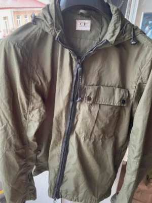Cp company goggle jacket - Snygg olivgrön vindjacka från C.P. Company med dragkedja, huva med ikoniska glasögon och bröstficka med knapp. Jackan har en avslappnad passform och är tillverkad i lätt material, perfekt för blåsiga dagar. Storlek M men passar både S och M