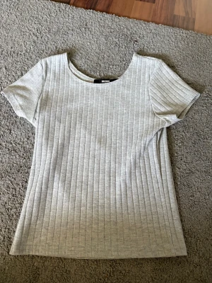 Ljusgrå ribbad topp från BikBok - Superenkel och stilren ljusgrå ribbad topp från BikBok. Toppen har korta ärmar, rund halsringning och är gjord i ett mjukt material som sitter skönt. Perfekt att matcha med jeans eller kjol för en clean look.