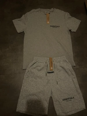 Grått loungewear-set från Essentials - Snyggt och bekvämt loungewear-set från Essentials i ljusgrått. Setet består av en t-shirt med rund hals och tryck på bröstet samt matchande shorts med dragsko och fickor. Perfekt för chill dagar hemma. Mjukt material som känns skönt mot huden.