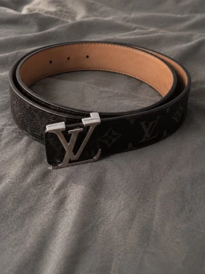  svart skärp med monogram -  skärp i svart skinn med klassiskt monogrammönster och stor logga i silver på spännet. Insidan är beige och bältet har en exklusiv känsla som passar till många olika stilar. Sparsamt använt 