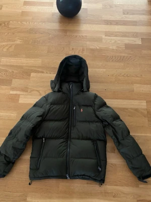 Mörkgrön pufferjacka från Polo Ralph Lauren - Snygg mörkgrön pufferjacka från Polo Ralph Lauren med huva och dragkedjor framtill. Jackan har en klassisk pufferdesign, orange logga på bröstet och flera praktiska fickor. Perfekt för kalla dagar när du vill hålla stilen.