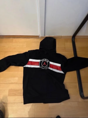 Svart PSG x Jordan hoodie - Svart hoodie med huva från Jordan x Paris Saint-Germain. Stor logga på bröstet med Paris Saint-Germain och Jumpman-symbolen, samt röda och vita ränder över bröstet. Perfekt för dig som gillar sportig streetstyle.
