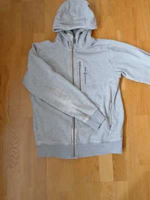 Ljusgrå hoodie från sail racing - Snygg ljusgrå hoodie från Peak Performance med dragkedja hela vägen och en praktisk bröstficka med dragkedja. Klassisk huva och ribbade muddar vid ärmslut och nederkant. Perfekt för en avslappnad och sportig stil. Sitter som en s/m.