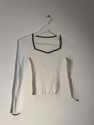 Vit ribbad långärmad topp - Vit ribbad långärmad topp med svart kant runt den fyrkantiga ringningen och ärmsluten. Toppens material är stretchigt och figurnära, perfekt för en trendig och stilren look.
