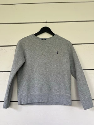 Grå sweatshirt från Polo Ralph Lauren - Klassisk grå sweatshirt från Polo Ralph Lauren med rund halsringning och broderad logga på bröstet. Tröjan har långa ärmar och ribbade muddar vid ärmslut och nederkant. Perfekt för en avslappnad och stilren look.