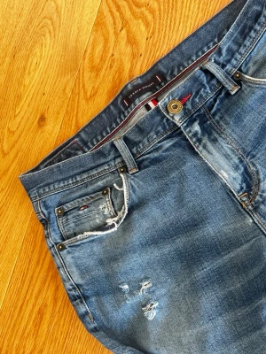 Blå slitna jeans Tommy Hilfiger - Säljer ett par blå jeans från Tommy Hilfiger med slitningar och distressed detaljer på både fram- och baksida. Klassisk femficksmodell med smal passform och snygga slitna kanter. Perfekta för en avslappnad och trendig look. W33 L32
