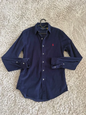 Marinblå manchesterskjorta Polo Ralph Lauren - Snygg marinblå manchesterskjorta från Polo Ralph Lauren i slim fit-modell. Skjortan har långa ärmar, klassisk krage, knäppning framtill och ikonisk röd logga broderad på bröstet. Perfekt för sommaren