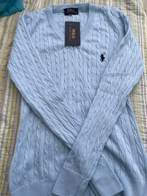 Ljusblå kabelstickad tröja Polo Ralph Lauren - Ljusblå kabelstickad tröja från Polo Ralph Lauren med V-ringning och klassisk broderad logga på bröstet. Tröjan har långa ärmar och ribbade muddar vid ärmslut och nederkant. Perfekt för en stilren och avslappnad look.