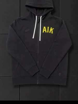 Nike hoodie aik - Svart hoodie från Nike med AIK-tryck i gult på bröstet. Tröjan har huva med vita snören och dragkedja framtill. Tillverkad i mjukt material, perfekt för en sportig och avslappnad stil. Praktiska fickor på sidorna och klassisk passform. Storlek S