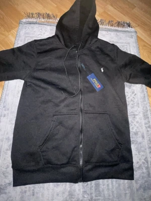 Svart hoodie från Polo Ralph Lauren - Snygg svart hoodie med dragkedja från Polo Ralph Lauren. Klassisk design med huva och dragsnören, två stora fickor framtill och den ikoniska lilla loggan broderad på bröstet. Perfekt för en avslappnad och stilren look.