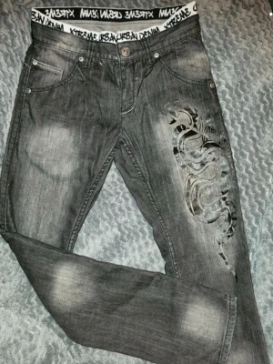 Snygga jeans  - Snygga grå jeans från kapphal i storlek 164 med coolt drakmönster på ena benet och tydliga slitningar. Jeansen har midjeresår med logga. Perfekta för dig som vill sticka ut med din stil. 