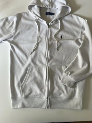 Vit hoodie från Polo Ralph Lauren - Snygg vit hoodie med dragkedja från Polo Ralph Lauren. Klassisk design med huva, dragsnören och kängurufickor. Liten broderad logga på bröstet. Mjuk bomullsblandning som är skön att bära. Perfekt för en clean och stilren look. Pris kan diskuteras!!!