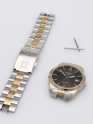 Tissot PR 100 herrklocka svart/guld - Stilren Tissot PR 100 herrklocka med svart urtavla, guldfärgade detaljer och datumvisning. Armbandet är i silver och guldtonat rostfritt stål. Klassisk rund form och tydliga indexmarkeringar. Perfekt accessoar för dig som gillar tidlös design.
