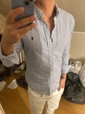 Randig skjorta från Ralph Lauren - Klassisk blåvit randig skjorta från Ralph Lauren med button-down krage och broderad logga på bröstet. Skjortan har lång ärm, knappar framtill och är tillverkad i bomull. Perfekt för en stilren look.