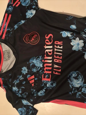 Real Madrid Adidas träningströja blommig - Snygg svart träningströja från Adidas och Real Madrid med röda detaljer och stora blå blommor. Tröjan har klubbmärke och sponsortryck framtill samt klassiska Adidas-ränder på axlarna. Perfekt för dig som vill sticka ut på planen. Storlek 154
