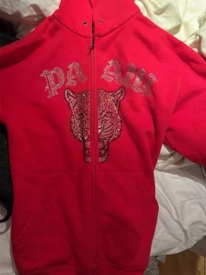 Röd PAWZ hoodie med glittertryck - Snygg röd hoodie med dragkedja från PAWZ. Framsidan har ett glittrigt tryck med texten 'PAWZ' och ett stort tigerhuvud i paljetter. Hoodien har huva och fickor framtill, perfekt för en cool och avslappnad stil.