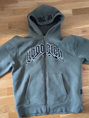 Olivgrön zip-hoodie från Hoodrich - Olivgrön hoodie med dragkedja från Hoodrich. Stor broderad logga på bröstet och tryck i huvan. Mjuk insida, känguruficka och klassisk streetwear-stil. Perfekt för dig som gillar snygga och bekväma plagg.