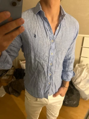 Ljusblå linneskjorta från Ralph Lauren - Snygg ljusblå skjorta i linne från Ralph Lauren med klassisk krage och knappar framtill. Skjortan har lång ärm och den ikoniska loggan broderad på bröstet. Perfekt för en avslappnad men stilren look.