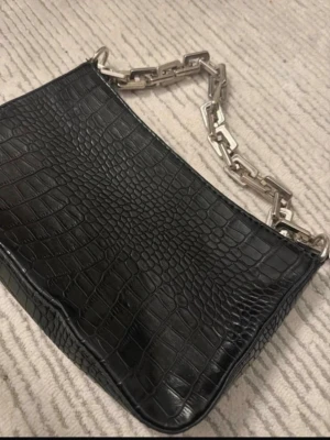 Svart croco-handväska med kedja - Stilren svart handväska i croco-mönstrat konstläder med en chunky silverfärgad kedja som axelrem. Väskan har en rektangulär form och ger en edgy vibe till din outfit. Perfekt för dig som gillar trendiga accessoarer.