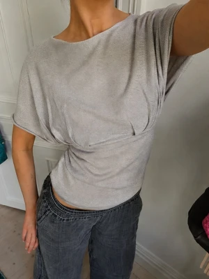 Glittrig silver topp med kort ärm - Snygg glittrig silver topp med kort ärm och lös passform. Toppen har en lätt draperad effekt vid midjan och en rund halsringning. Perfekt för dig som vill sticka ut lite extra med en stilren look. SÅ HIMLA SNYGG !!!!!!! Hittade 2nd hand så går inte att köpa i butik 🩷