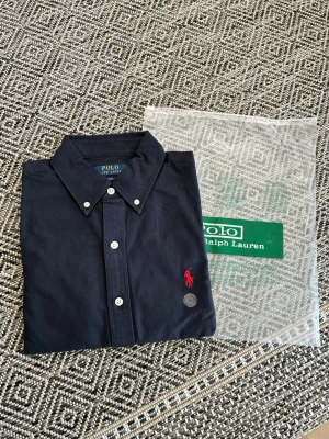 Marinblå skjorta från Polo Ralph Lauren - Klassisk marinblå skjorta från Polo Ralph Lauren med röd broderad logga på bröstet. Skjortan har knappar framtill, button-down krage och långa ärmar. Tillverkad i mjuk bomull som känns skön mot huden. Perfekt för en stilren och avslappnad look.