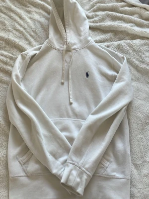 Vit hoodie från Polo Ralph Lauren - Vit hoodie från Polo Ralph Lauren med klassisk huva och dragsko. Broderad marinblå logga på bröstet och stor känguruficka framtill. Mjuk bomullskvalitet och avslappnad passform, perfekt för en chill och stilren look. Finns tecken på användning men inte något som man verkligen märker