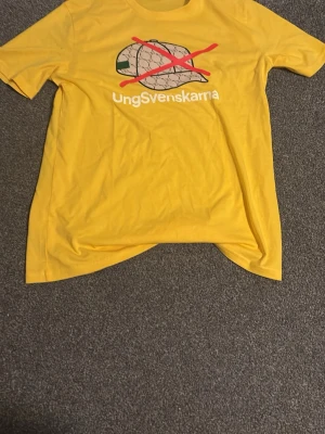 Gul t-shirt Ungsvenskarna - Gul t-shirt från Ungsvenskarna med tryck av en keps med rött kryss och texten 'Ungsvenskarna' på bröstet. Klassisk passform med rund hals och korta ärmar. Perfekt för dig som vill sticka ut med ett statement-plagg.
