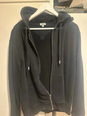 Svart Kenzo hoodie med dragkedja - Svart hoodie från Kenzo med dragkedja framtill och stora fickor. Huva med dragsko och vit Kenzo Paris-logga tryckt på huvan. Mjuk bomullskänsla och klassisk passform, perfekt för en chill och stilren look. Storlek L men krympt till M