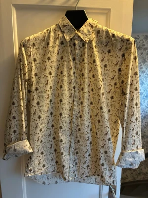 Vit skjorta med djurmönster Robbie Moor - Vit skjorta från Robbie Moor med ett unikt djurmönster som inkluderar giraffer och palmer. Skjortan har lång ärm, klassisk krage och knäppning framtill. Slim fit-modell som ger en snygg och modern passform. Perfekt för dig som vill sticka ut lite extra.