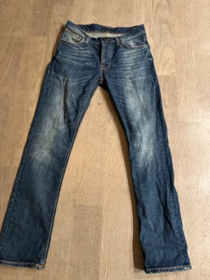 Blå jeans från Nudie Jeans - Snygga blå jeans från Nudie Jeans med klassisk femficksdesign och orangea sömmar. Jeansen har en rak passform och slitningar framtill för en cool, använd look. Tillverkade i denim med detaljerade bakfickor och läderpatch i midjan.