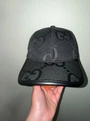 Gucci keps  - Snygg svart keps från Gucci med det klassiska GG-mönstret i mörkgrått. Kepsen är tillverkad i bomull och har en böjd skärm med läderdetalj runt kanten. Perfekt accessoar för en trendig och exklusiv look. 