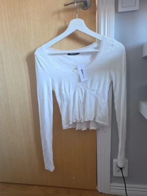 Vit spetsdetaljerad topp från gina tricot - Supersöt vit topp från gina tricot med spetsdetaljer vid halsringningen och framtill. Modellen är croppad med lång ärm och har en lätt volang nertill. Perfekt att styla med jeans eller kjol för en trendig look.