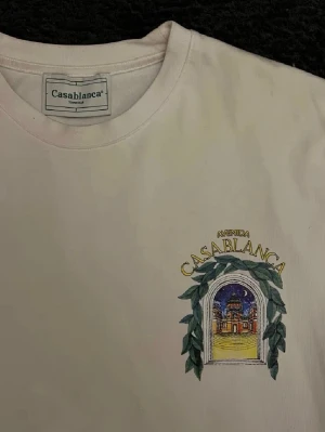 Vit t-shirt från Casablanca - Vit t-shirt från Casablanca med tryck på bröstet som föreställer en färgglad byggnad och gröna blad. Klassisk passform och rund halsringning. Perfekt för dig som gillar stilrena plagg med unika detaljer.