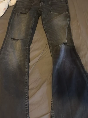 Svarta slitna jeans med stora fickor - Säljer ett par svarta jeans med slitningar och hål på båda benen. Jeansen har stora framfickor och en cool, avslappnad look. Perfekta för dig som gillar streetstyle och vill ha något unikt i garderoben.