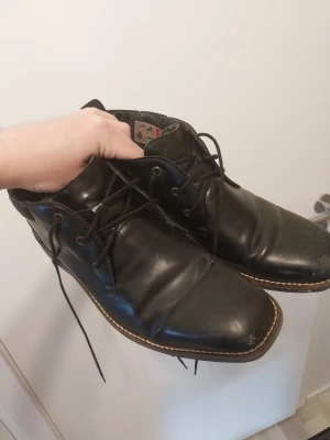 Svarta läderloafers med snörning Rieker - Säljer ett par svarta loafers i läder med snörning och kontrastsöm längs sulan. Skorna har en klassisk, något fyrkantig tå och stilren design. Perfekta för dig som gillar en clean och tidlös look.