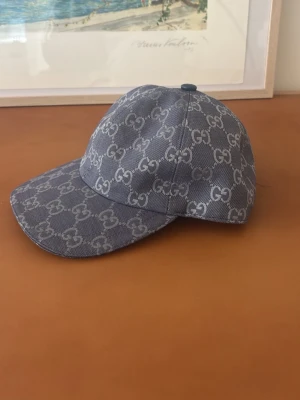 Blå keps från Gucci med GG-mönster - Snygg blå keps från Gucci i denim-look med det klassiska GG-monogrammet över hela kepsen. Böjd skärm och justerbar baksida. Perfekt för dig som vill sticka ut med en exklusiv accessoar.