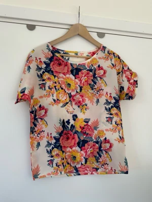Blommig topp med kort ärm - storlek M - Härlig blommig topp / blus från Liva Girl med korta uppvikta ärmar och rund halsringning. Storlek M. Vackert mönster av stora blommor i olika färger som rosa, gult och blått på en ljus bakgrund. Enkel att matcha med både byxor och kjol. Mycket gott skick, som ny!