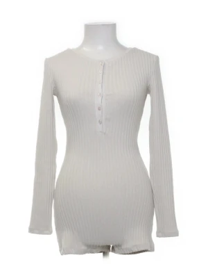 Vit ribbad playsuit med lång ärm - Säljer en stilren vit body med lång ärm och ribbad struktur. Bodyn har rund halsringning och knappar framtill för en snygg detalj. Tillverkad i mjukt, stretchigt material som sitter skönt på kroppen. Perfekt för dig som gillar enkel och clean stil.