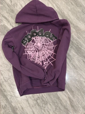 Lila Sp5der hoodie med spindelnät - Cool lila hoodie från Sp5der med stor rosa spindelnätsgrafik och glittrande text på ryggen. Hoodien har huva och en avslappnad passform, perfekt för en streetwear-look. Materialet är mjukt och bekvämt, passar dig som gillar unika detaljer.