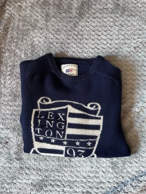 Mörkblå stickad tröja från Lexington - Mörkblå stickad tröja från Lexington med stort vitt emblem och text framtill. Tröjan har rund halsringning och ribbade muddar vid ärmslut och nederkant. Perfekt för dig som gillar klassisk och stilren design.
