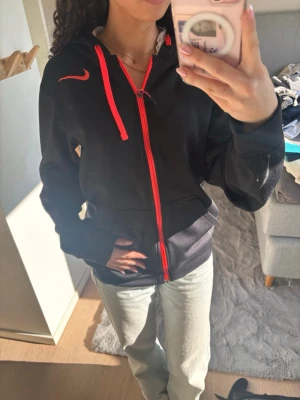 Svart Nike hoodie med röd dragkedja - Svart hoodie från Nike med röd dragkedja och röda snören i huvan. Klassisk Nike-logga i rött på bröstet. Hoodien har fickor framtill och är tillverkad i mjukt material som känns skönt mot huden. Perfekt för en sportig och avslappnad stil.