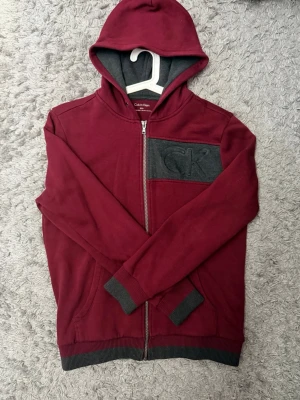 Vinröd hoodie från Calvin Klein - Snygg vinröd hoodie från Calvin Klein med dragkedja och huva. Gråa detaljer på muddar, fickor och insida av huvan samt en grå bröstficka med CK-logga. Perfekt för en chill och stilren look. Sitter rätt så baggy. Bara att skriva vid minsta fundering