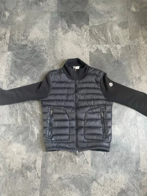 Svart pufferjacka med dragkedja moncler cardigain - Snygg svart pufferjacka med quiltat mönster och två dragkedjefickor framtill. Jackan har svarta ärmar i mjukt material och hög krage. Perfekt för kalla dagar och har en stilren look med diskret logga på ärmen.